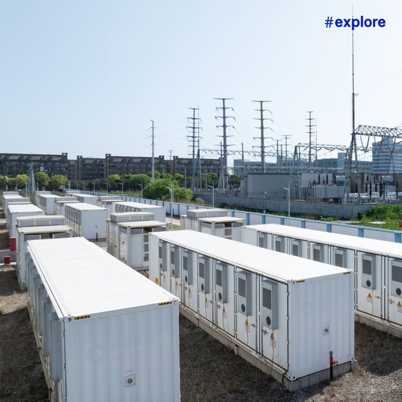 Energieanlage mit Transformatoren Eine industrielle Energieanlage mit mehreren weißen Containergebäuden und Transformatoren. Im Hintergrund sind Strommasten und ein Gebäude zu sehen. Die Anlage ist von einer grünen Landschaft umgeben und der Himmel ist klar und blau.