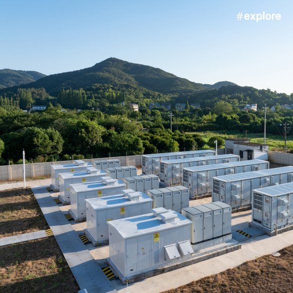 Energieanlage in grüner Umgebung Eine moderne Energieanlage mit mehreren weißen Containergebäuden, die in einer grünen Landschaft mit Hügeln im Hintergrund liegt. Der Himmel ist klar und blau, und der Hashtag "#explore" ist oben rechts im Bild zu sehen.