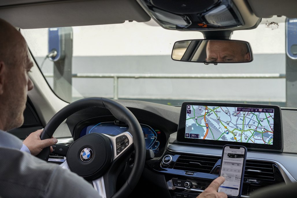 Fahrer in einem BMW-Fahrzeug mit Navigationssystem auf dem Display und einem Smartphone in der Hand, sichtbar im Rückspiegel.