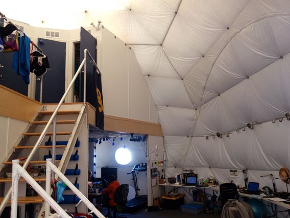 Innenraum einer weißen Kuppelstruktur mit Treppe, Arbeitsplatz und hängenden weißen Stoffbahnen an der Decke.