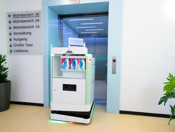 Ein weißer Transportroboter mit grüner LED-Beleuchtung steht vor einem Aufzug in einem modernen Gebäude mit deutschsprachiger Beschilderung.