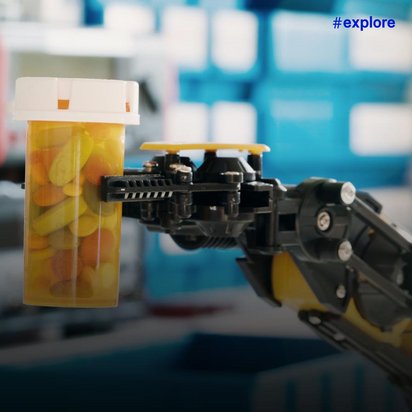 Ein schwarzer Roboterarm greift präzise nach einer gelben Medikamentenschachtel in einer technischen Umgebung.