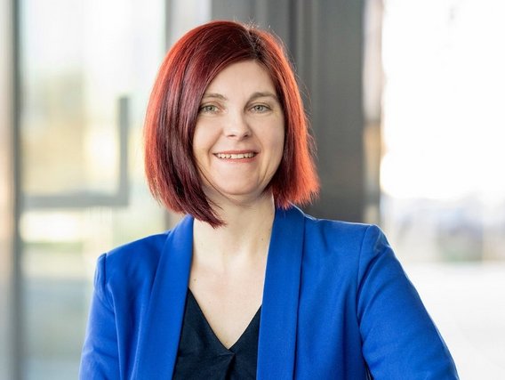 Dr. Nele Pollmann mit rotbraunen schulterlagen Haaren in einer blauen Blazerjacke, die in einem modernen Büroumfeld steht.