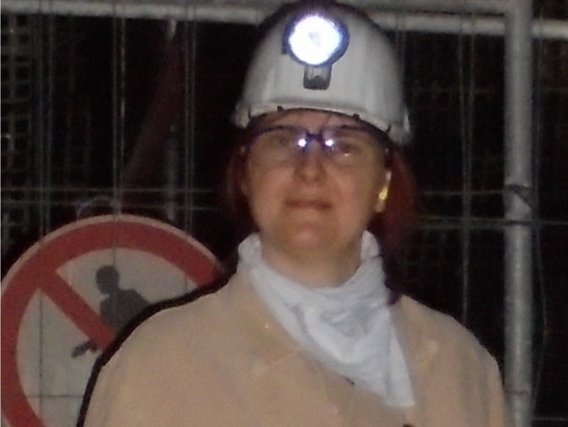 Nele Pollmann mit Schutzhelm und Sicherheitsausrüstung in einem industriellen Umfeld, mit einem Warnschild im Hintergrund.