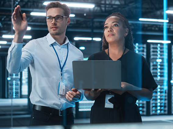 Two people are standing in a modern server room. One person is wearing a white shirt and a blue ID card around their neck, while the other is wearing a black top and holding a laptop. Several illuminated server racks can be seen in the background.
