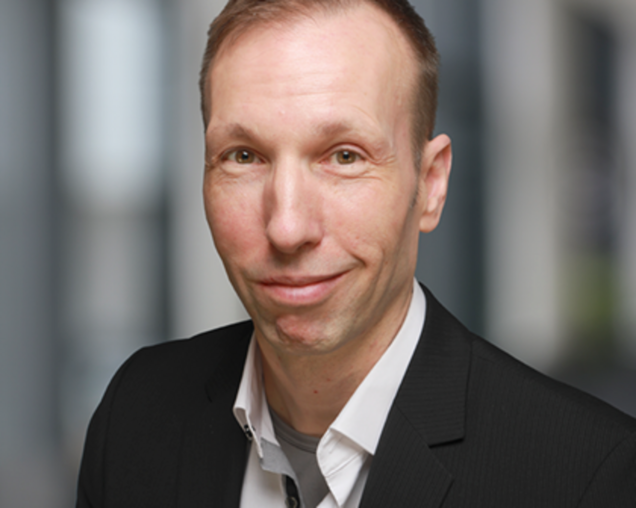 Dietmar Rosenthal in schwarzem Anzug und weißem Hemd mit kurzem Haar und lächelndem Gesicht vor einem unscharfen Hintergrund.