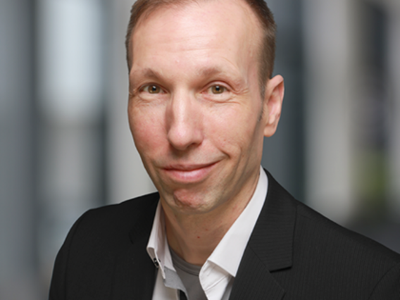 Dietmar Rosenthal in schwarzem Anzug und weißem Hemd mit kurzem Haar und lächelndem Gesicht vor einem unscharfen Hintergrund.