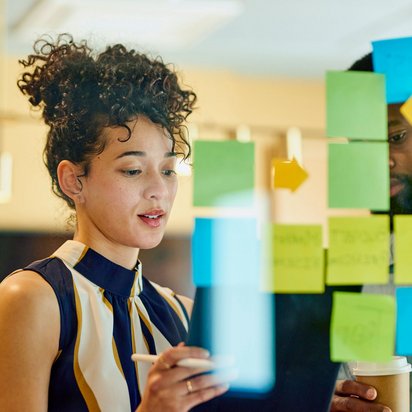Kreative Teamarbeit im Projektmanagement Frau steht vor einer Glaswand mit bunten Haftnotizen und spricht über ein Projekt.