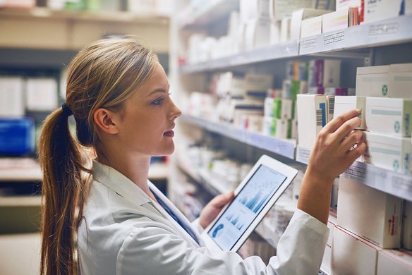 Apothekerin prüft Medikamente mit digitaler Unterstützung im Apothekenregal Apothekerin mit Tablet entnimmt Medikamentenschachtel aus einem Regal in der Apotheke.