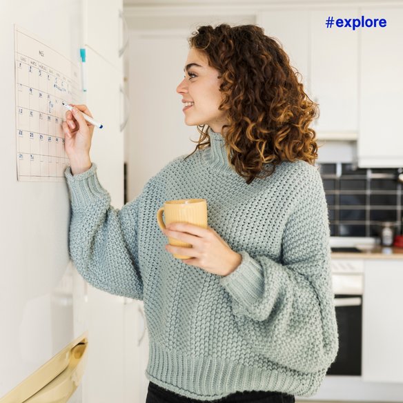Planung im Alltag Eine Person steht in einer Küche und markiert ein Datum auf einem Wandkalender. Sie trägt einen gemütlichen Strickpullover und hält eine gelbe Tasse in der Hand. Der Hashtag "#explore" ist oben rechts im Bild zu sehen.