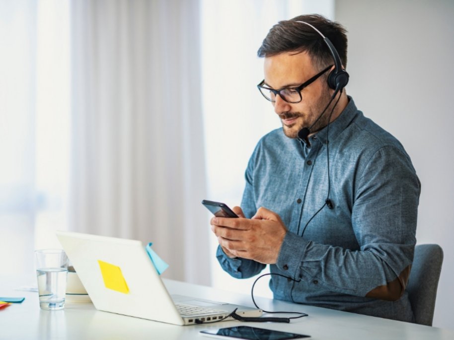 Ein Mann mit Brille und Headset in einem blauen Hemd schaut auf sein Smartphone, während er an einem Tisch mit Laptop und Wasserglas sitzt.