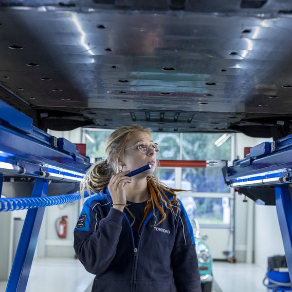 Fahrzeuguntersuchung auf der Hebebühne Technikerin bei der Untersuchung eines angehobenen Fahrzeugs in einer Werkstatt. Im Hintergrund steht ein weiteres Auto.