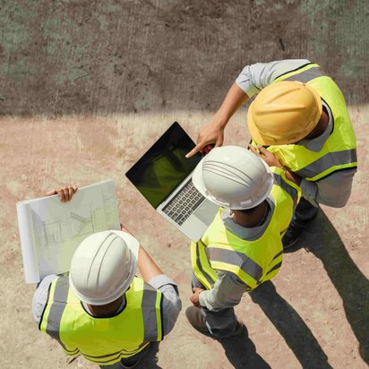 Drei Bauingenieure in Sicherheitswesten und Helmen stehen auf einer Baustelle und betrachten gemeinsam einen Laptop und Baupläne.