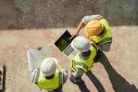 Drei Bauingenieure in Sicherheitswesten und Helmen stehen auf einer Baustelle und betrachten gemeinsam einen Laptop und Baupläne.