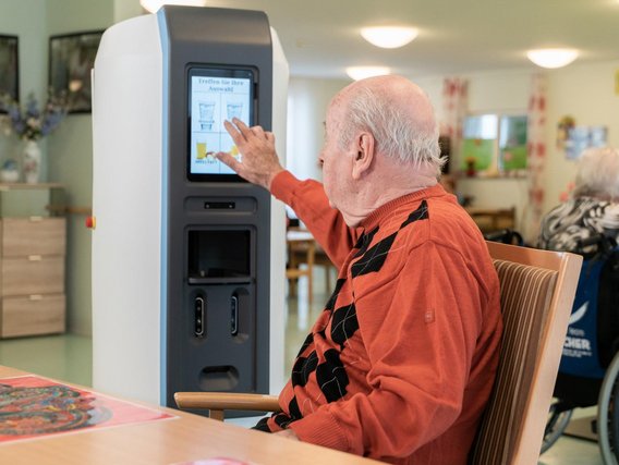 Ein älterer Mann im orangefarbenen Pullover bedient einen Touchscreen-Automaten in einem hellen Gemeinschaftsraum.