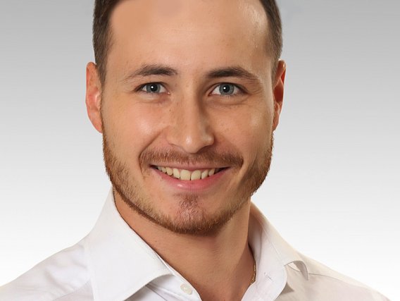 Denis Raschka in einem weißen Hemd lächelt freundlich und selbstbewusst.