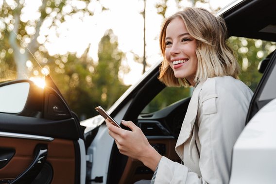 MPU-Gutachten bei TÜV NORD – alle Infos Lächelnde junge Frau sitzt mit Smartphone in der Hand in der offenen Tür eines Autos