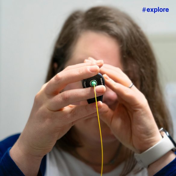 Person hält ein gelbes Kabel und betrachtet es durch ein Gerät Eine Person hält ein gelbes Kabel und betrachtet es durch ein Gerät. Die Person trägt ein weißes Armband und hat braunes Haar. Oben rechts im Bild ist der Hashtag "#explore" in blauer Schrift zu sehen.