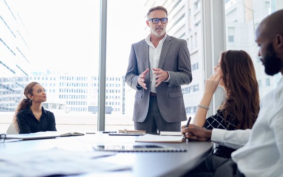 Präsentation eines Projektleiters im Business-Meeting. Mann im Anzug spricht vor einer Gruppe von Personen in einem modernen Konferenzraum.