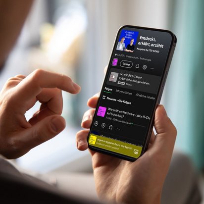 Person hält ein Smartphone in der Hand, auf dem eine Podcast‑Übersichtsseite mit Episodenliste angezeigt wird. Der Bildschirm zeigt verschiedene Folgen sowie Bedienelemente einer Audio‑App. Der Hintergrund ist unscharf gehalten.