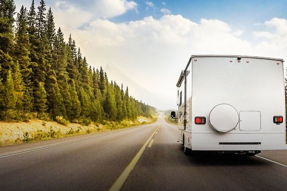 Ein weißes Wohnmobil fährt auf einer zweispurigen Straße durch eine bewaldete Landschaft mit Nadelbäumen. Der Himmel zeigt eine Mischung aus Wolken und blauem Himmel, während die Straße schnurgerade in die Ferne führt.