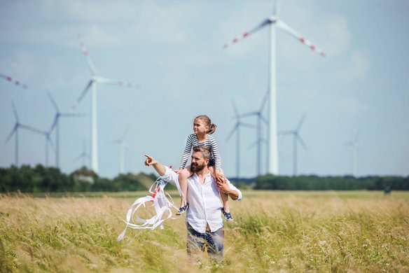 A man carries a child on his shoulders. They are standing in a green meadow with tall grass, and several wind turbines can be seen in the background.