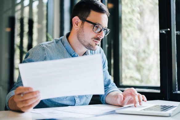 Ein konzentrierter Mann prüft ein Papierdokument und gibt Daten in seinen Laptop ein. Er sitzt an einem modernen Arbeitsplatz am Fenster. Analytisches Arbeiten im modernen Büro
