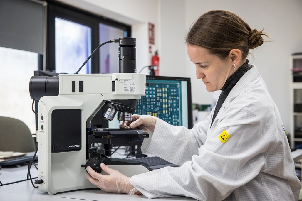 Wissenschaftliche Untersuchung am Mikroskop Eine Person in einem Laborkittel arbeitet mit einem Mikroskop in einem Labor. Im Hintergrund ist ein Computerbildschirm mit technischen Diagrammen zu sehen. Die Person trägt Handschuhe und konzentriert sich auf die Analyse.