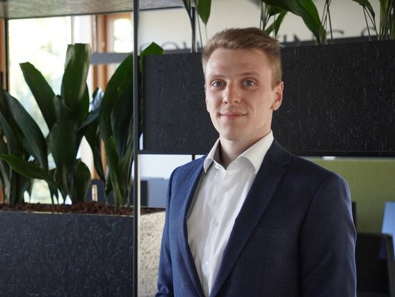 Jan Simoneit in blauem Anzug und weißem Hemd steht vor einer schwarzen Trennwand mit Zimmerpflanzen in einem modernen Büroraum.