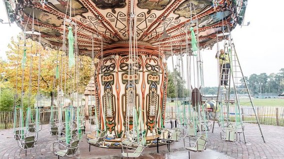 Ein traditionelles Kettenkarussell mit kunstvoll verzierter Mitte und leeren Sitzen, aufgestellt auf gepflastertem Boden in einem Freizeitpark im Herbst.