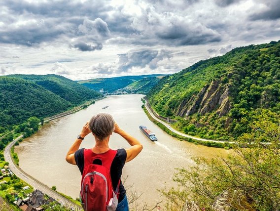 Eine Wanderin mit rotem Rucksack blickt auf einen gewundenen Fluss (vermutlich den Rhein) durch ein grünes Tal mit bewaldeten Hügeln und Felswänden, während ein Frachtschiff auf dem Wasser fährt und dramatische Gewitterwolken den Himmel bedecken.