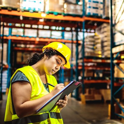 Lagerlogistik und Bestandsmanagement im Überblick Mitarbeiterin mit Schutzweste und Helm dokumentiert Lagerbestände in einem großen Hochregallager