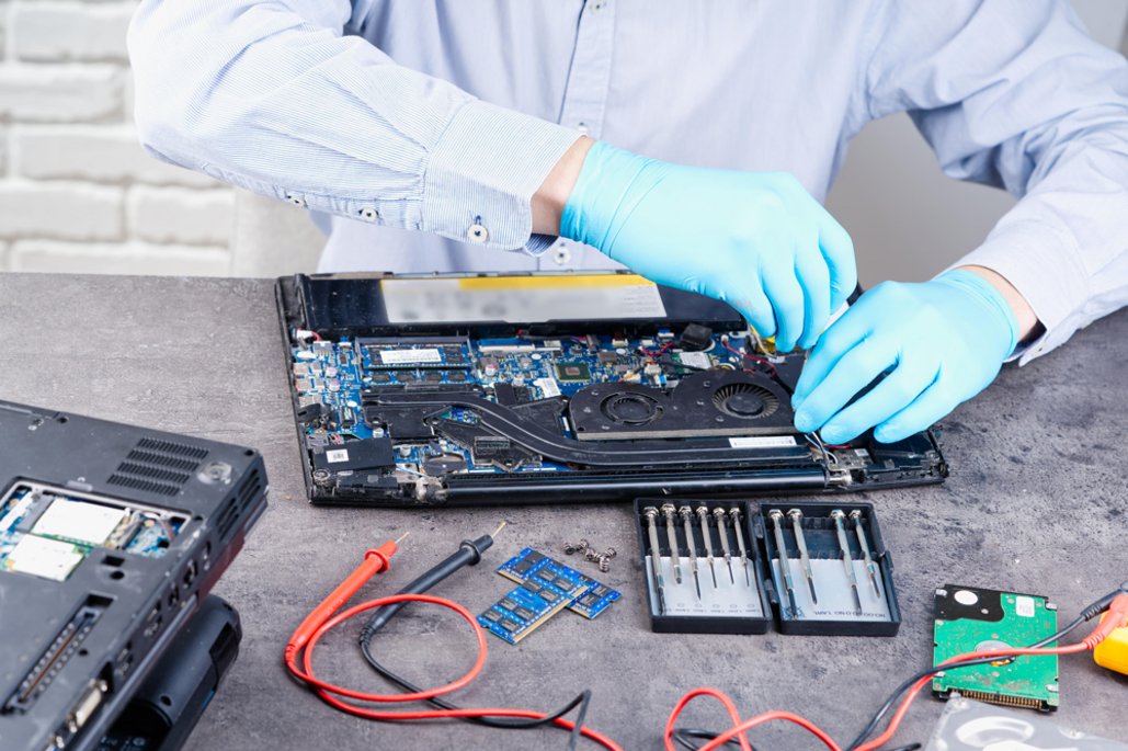 Eine Person mit blauen Handschuhen repariert einen Laptop, dessen Inneres geöffnet ist. Verschiedene Werkzeuge und Komponenten liegen auf dem Tisch.