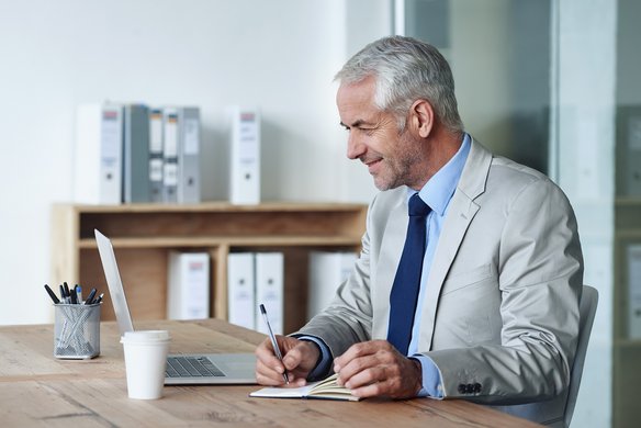 Professionelles Arbeiten im digitalen Büro. Älterer Mann im Anzug sitzt am Schreibtisch, lächelt und arbeitet mit Laptop und Notizblock.