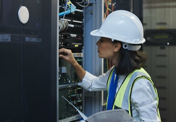 IT-Inspektion: Fachgerechte Wartung im Rechenzentrum. Technikerin mit Helm prüft Serverkomponenten in einem Rechenzentrum.