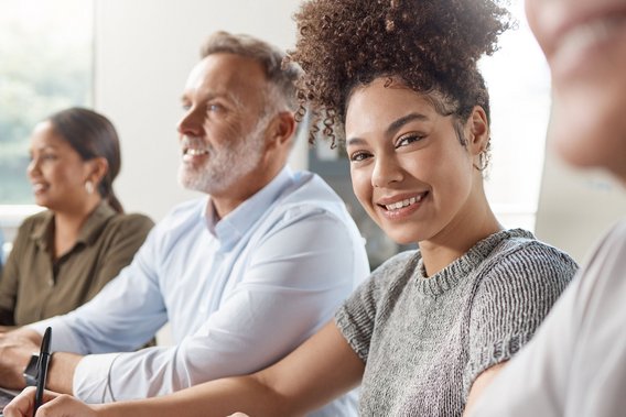Startseite von TÜV NORD Transfer – Bildung. Beratung. Integration. Lächelnde junge Frau mit Stift in der Hand sitzt mit anderen Personen in einem Seminarraum