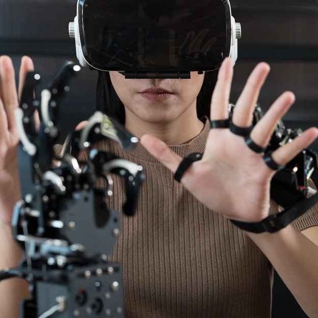 Frau mit KI-Technologie Eine Person mit VR- oder AR-Brille interagiert mit erhobenen Händen mit einer Roboterhand.