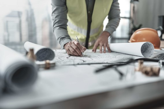 Person in Sicherheitsweste arbeitet an Bauplänen auf einem Tisch mit Bauhelm und Werkzeugen.