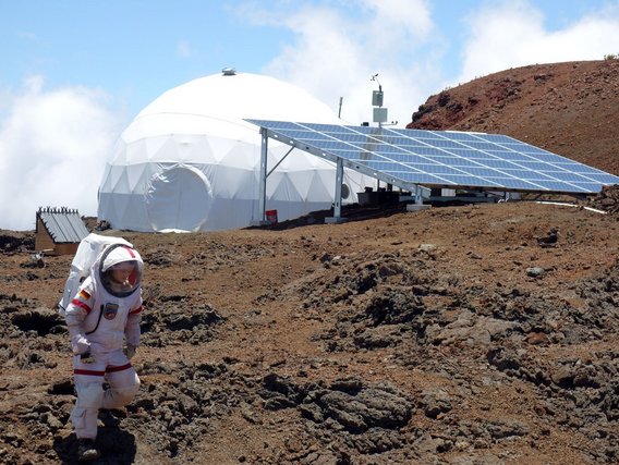Person in weißem Raumanzug vor weißer Kuppelstation mit Solarpaneelen in rötlicher Vulkanlandschaft.