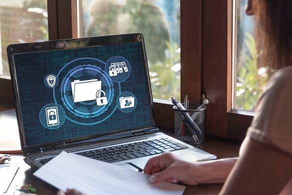 DSGVO und Bußgelder: Datenschutzverstöße erkennen und vermeiden. | TÜV Laptop mit Datenschutzsymbolen auf dem Bildschirm und Person mit Dokument am Schreibtisch.