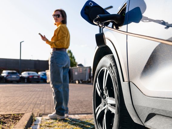 TÜV NORD-Ladesäule Frau steht neben einem Elektroauto an einer Ladestation und nutzt ihr Smartphone.