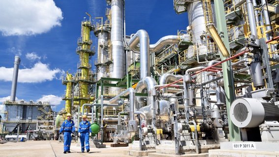 TÜV NORD Dampf- und Drucktechnik Zwei Männer in blauen Arbeitsanzügen und mit weißen Schutzhelmen laufen nebeneinander vor dem Hintergrund eines Chemieparks. Es sind diverse technische Anlagen unter blauen Himmel zu sehen.