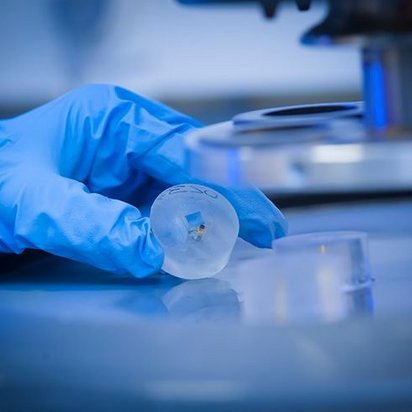 Eine Hand in einem Handschuh hält eine Kugel aus Kunststoff vor einem Mikroskop