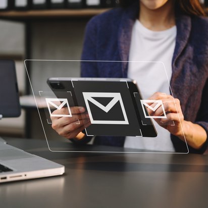 Person hält Tablet mit schwebenden E-Mail-Symbolen, Laptop und Kaffeebecher im Hintergrund.