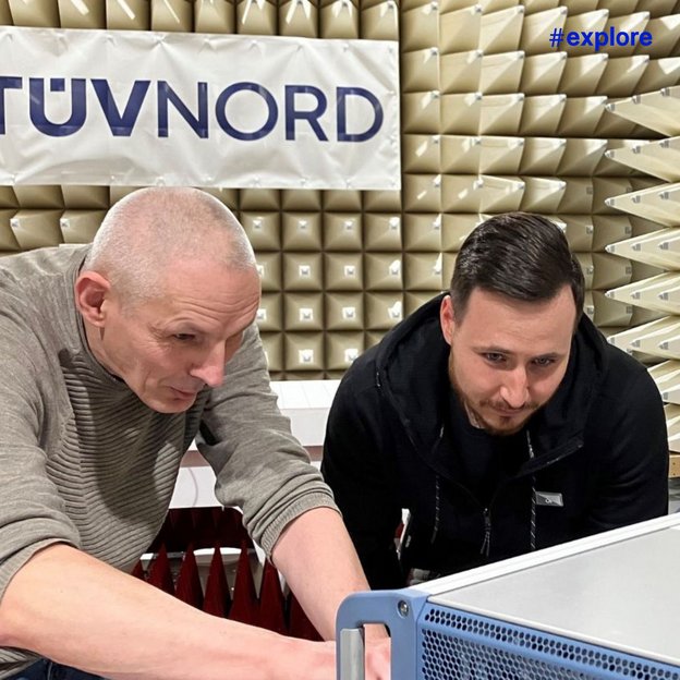 Zwei Männer arbeiten konzentriert an einem technischen Gerät in einem TÜV Nord Prüflabor mit Absorptionspyramiden im Hintergrund.