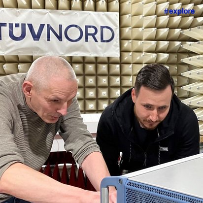 Zwei Männer arbeiten konzentriert an einem technischen Gerät in einem TÜV Nord Prüflabor mit Absorptionspyramiden im Hintergrund.