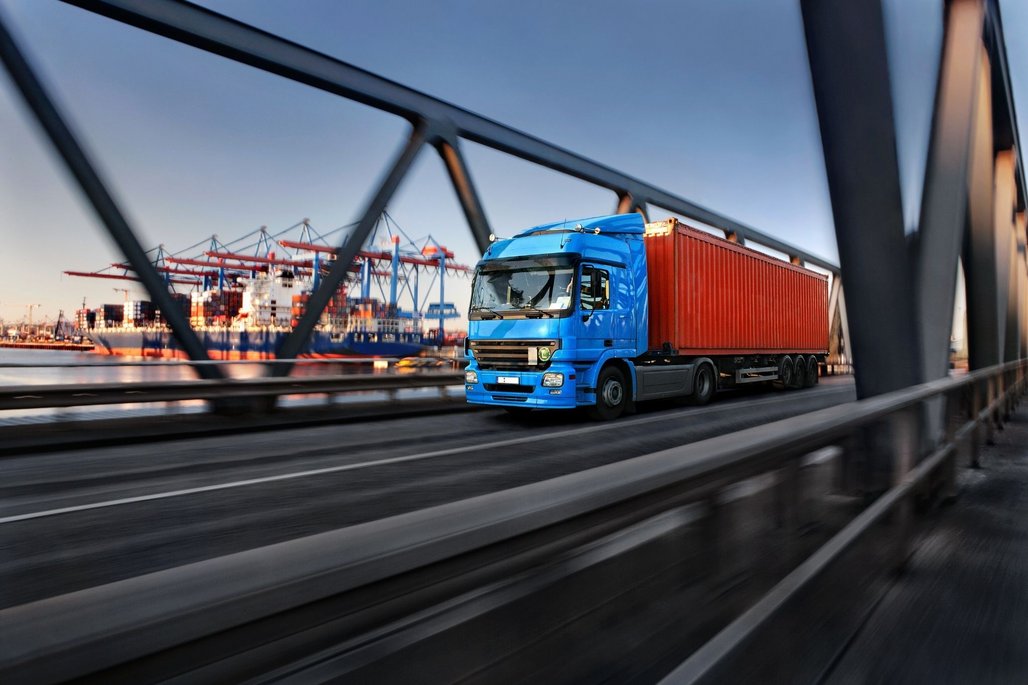 LKW auf Brücke nahe Hafen Ein blauer LKW transportiert einen roten Container über eine Brücke. Im Hintergrund ist ein Hafen mit Containerschiffen und Kränen zu sehen. Die Szene vermittelt Bewegung und industrielle Aktivität.