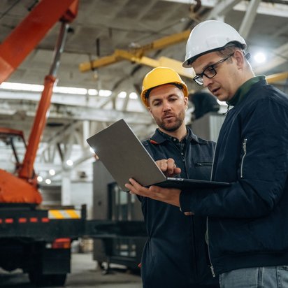 Digitale Kontrolle industrieller Prozesse vor Ort Zwei Ingenieure mit Schutzhelmen prüfen Produktionsdaten auf einem Laptop in der Werkshalle