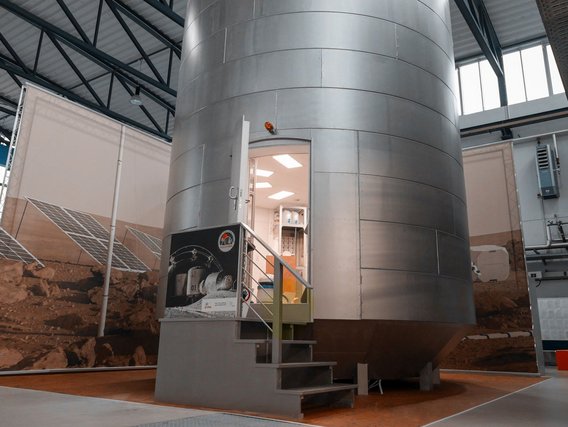 Großer silberner zylindrischer Simulator in einer Industriehalle mit Treppe zum Eingang und Solarpaneelen im Hintergrund.