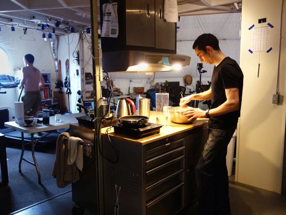 Mann in schwarzem Shirt kocht in einer kompakten Küche, im Hintergrund eine weitere Person an einem Laufband.